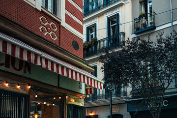 Obraz premium A charming building in Madrid featuring striped awnings, iron balconies, and warm evening lights in a lively neighborhood.