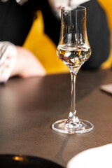Close-up of a clear stemmed glass with transparent liquid on a dark surface, blurred background with warm tones, minimalistic composition, elegant drinkware in casual setting.