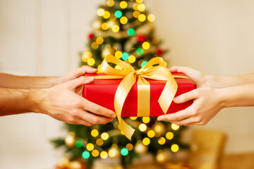 Hands Holding Red Christmas Gift With Golden Ribbon Against Festive Christmas Tree. Concept of Winter Holiday Celebration, Family Traditions, Seasonal Giving Spirit, And Christmas Visuals