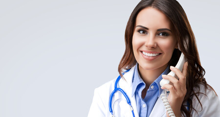 Portrait image of happy smiling cute female doctor on phone, isolated  against grey gray wall background with free text space. Medical call center concept.