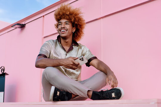 Relaxed African American man with curly ginger hair sitting outdoors holding smartphone, glancing sideways with smile, representing positive attitude toward mobile technology and connectivity.