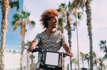 Young man with curly afro cycling shared rental bike with smile. Conceptual photo of eco-friendly mobility, app-based commuting, and integration of technology into daily lifestyle.