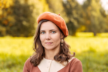 Headshot of a woman wearing an orange beret and vintage earrings standing in an autumn meadow