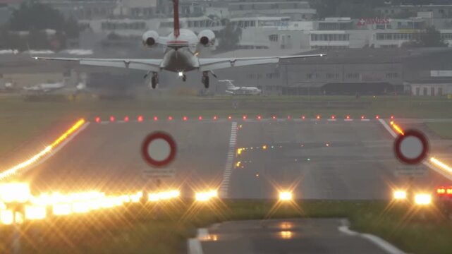 Small, private jet plane landing on an illuminated airport runway. Ground level, rear of the plane point of view, touchdown and taxi, real time, super-telephoto, cloudy, no people