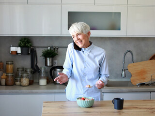 Portrait of a mid aged mature woman gettyng ready for work using a smartphone and eating breakfast oatmeal in a bowl standing in kitchen home. Overworked, busy, stress, urgency, multitasking, active h
