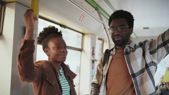 African American woman in striped shirt talking while holding handle on bus. Beside stands man with glasses listening closely. Riding together on public transportation. Conversation during commute