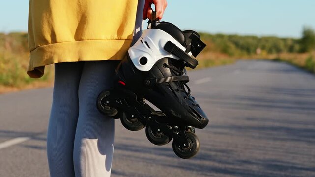 Bright sunlight shines on a young person holding black rollerblades by a serene road surrounded by green landscapes, perfect for skating