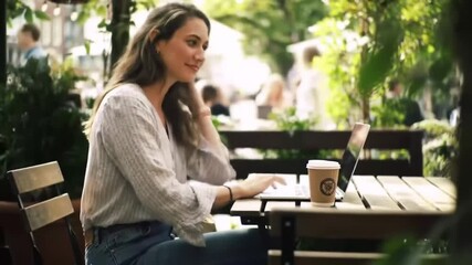 Happy young woman enjoying remote work on her laptop at an inviting outdoor cafe, sipping coffee amidst the vibrant urban atmosphere, embodying a flexible lifestyle - Powered by Adobe