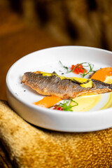 Gourmet plated fish dish with crispy skin, yellow sauce, orange puree, cherry tomatoes, and microgreens on a white ceramic plate over a warm textured background