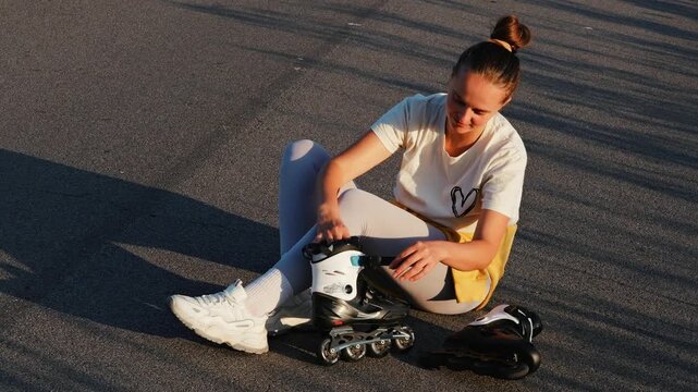A young woman sits on the pavement in soft light, tightening her rollerblades with a relaxed smile, ready for an afternoon skate