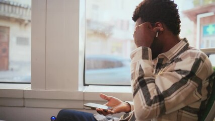 African American man wearing checkered shirt sitting by bus window putting earphones in ears. Passenger holds smartphone in hand preparing to listen music or podcast during ride. Public transport.