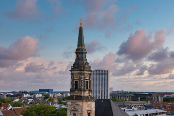 Fototapeta premium The spire of a historic church in Copenhagen, Denmark, stands out against a pastel sunset sky, surrounded by urban buildings and lush greenery.