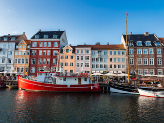 Fototapeta premium The Nyhavn district in Copenhagen, Denmark, features colorful historic buildings, a red boat in the canal, smaller vessels, and outdoor cafes under a clear sky.