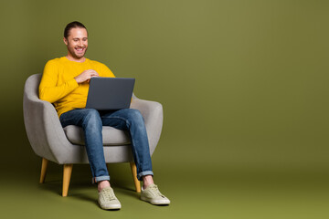 Young man in yellow sweater using laptop in a gray chair on a green background smiling and enjoying leisure time