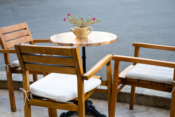 Charming outdoor café setup with wooden chairs, white cushions, and a teapot flower vase on a round table.