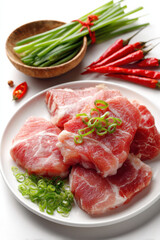 Fresh pork slices arranged on a plate with chopped green onions and red chili peppers displayed on a clean surface