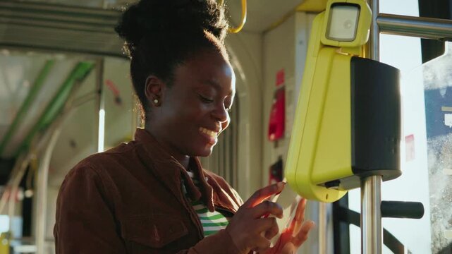 Charming African American woman holding smartphone close to yellow validator paying for ride on bus. Passenger in striped shirt and jacket completing contactless payment. Modern technology.