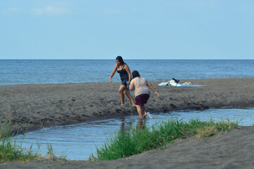 Carrying a drink and snacks, the mother and daughter walk together out of the shallow stream toward the sandy beach, enjoying their day near the tropical shoreline.