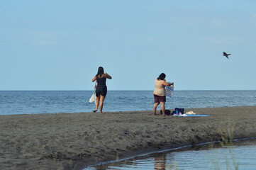 A teenage daughter walks barefoot on the sand carrying her dry outer dress, while her mother stands nearby adjusting her towel beside their mat near the brackish water.