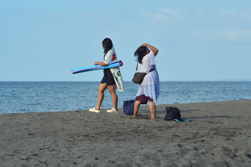 A daughter folds the blue beach mat while her mother fixes her wet hair, both standing on the sandy shoreline with belongings already packed, ready to leave peacefully.