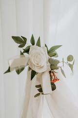 A beautifully arranged bunch of artificial flowers, including a white rose, is attached to a curtain. Green leaves and ribbon add an elegant touch to the interior decor.