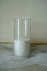 A tall, clear glass is filled with a white liquid sitting on a soft, light fabric. The background is neutral, creating a calm and simple atmosphere.