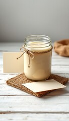 Creamy treat in rustic jar, jute twine detail, atop woven mat with blank card on textured wooden surface