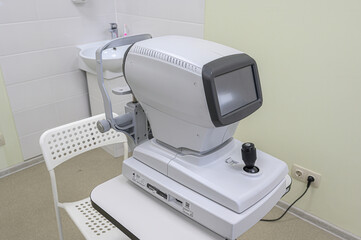A white autorefractor device is placed on a table with a white chair nearby. The device features a screen and adjustment knobs, set against a background of a sink and tiled wall