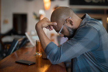 Senior Indian man sitting at wooden counter in pub with smartphone and half-full pint, copy space