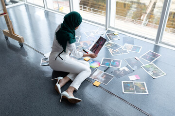 Middle Eastern hijabi woman sitting on studio floor, reviewing prints, color wheel and sticky notes