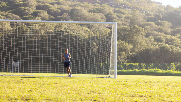 Boy guarding metal soccer goal frame inside net on sunlit grassy pitch by wooded hillside