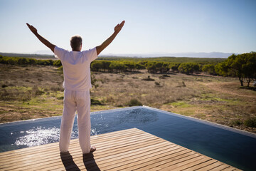 Middle-aged male stretching arms out on wooden pool deck by infinity pool overlooking scrub plain
