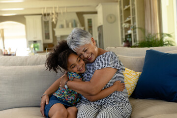 Snuggling senior Hispanic woman and African American girl on sofa in living room with throw pillows