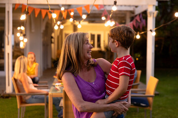 Multi-generational family gathering at table under pergola with lit string lights and bunting
