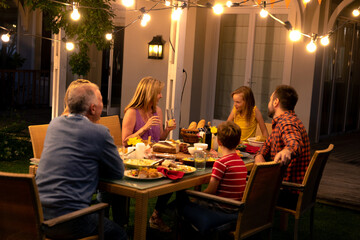 Multi generation family chatting around wicker dining table on backyard patio under string lights