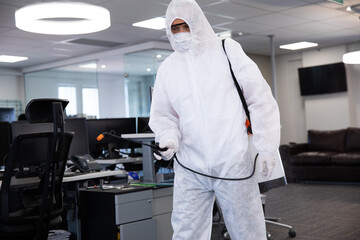 Male cleaning technician wearing coverall, spraying sanitizer across office with backpack sprayer