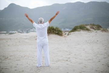 Senior man standing on wide sandy shoreline near dunes facing coastal town under overcast sky