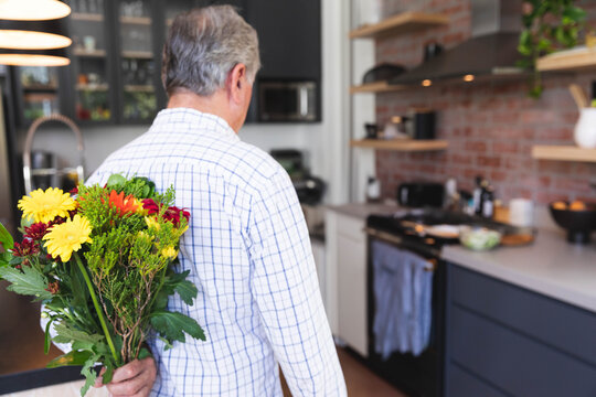 Senior man holding vibrant bouquet behind back in kitchen with brick backsplash and pendant lights - Powered by Adobe