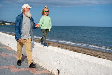 Smiling grandfather walking by the seaside promenade holding hands with his blond young grandson in green sweatshirt and sunglasses, as the boy balances on a low wall near the beach