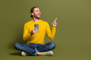 Attractive young man sitting cross-legged holding smartphone and pointing up on khaki background,...