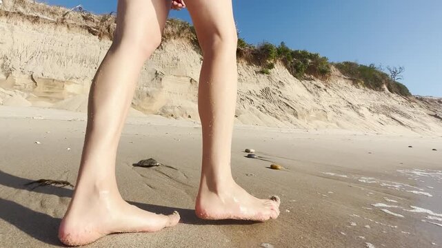 Close up of a woman walking barefoot on sandy beach near rocks and dunes. Steps in wet sand. Summer vacation, relaxation, freedom, seaside lifestyle, and nature journey
