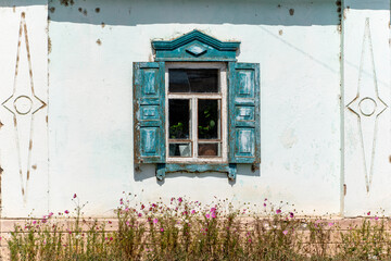 An old, weathered wooden window with peeling blue paint and shutters on a light-colored, textured wall of a traditional house.