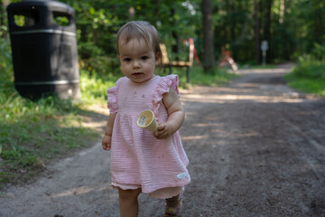 young toddler in pink dress exploring park path with ice cream cone, surrounded by lush greenery and gentle sunlight on a warm day, casual, playground, pastel, refreshment, environment, carefree