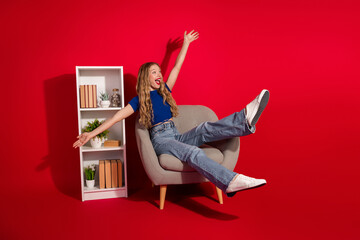 Joyful young woman on armchair against red background with bookshelf as she poses with outstretched...