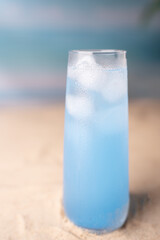 chilled blue refreshing drink in a tall glass set on sand with ocean background, creating a serene and cool beach vibe on a sunny day, freedom, exotic, therapeutic, luxury, escape, nature, idyllic