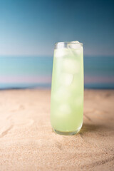 refreshing green beverage on sandy beach with ice cubes in tall glass against serene blue ocean horizon and soft gradient sky creating a relaxing summer ambiance, escape, soothing, harmony
