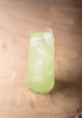 tall clear glass filled with refreshing light green beverage and ice cubes on softly textured wooden surface, evoking coolness and freshness in a minimalistic composition, neutral, peaceful, quiet
