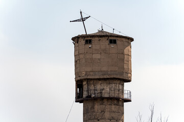 An old, weathered concrete water tower stands against a bright sky, featuring a worn, textured surface and small windows near its top.