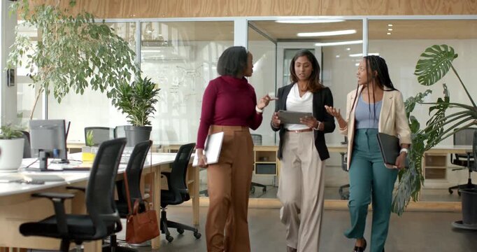 Diverse female colleagues in office walking, collaborating while reviewing tablet with white folder