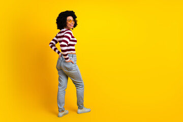 Fototapeta premium Young stylish woman with curly hair wearing striped pullover and jeans posing confidently against bright yellow background